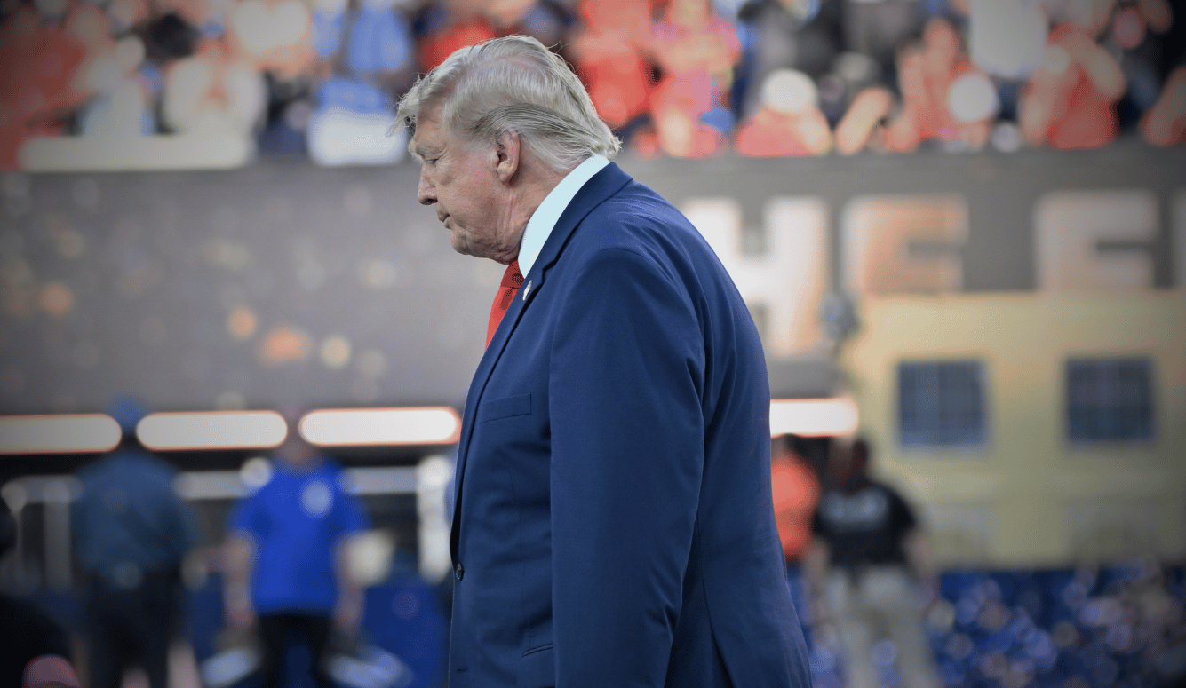 Trumpin a navy suit and red tie walking past a crowd at a stadium.