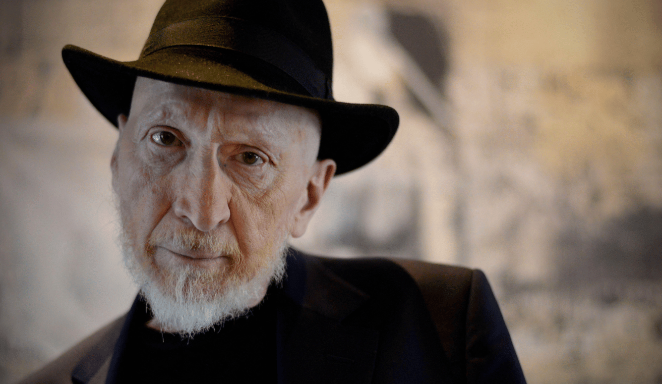 Frank Miller, with a white beard and black hat, looks towards the camera wearing a dark jacket and black shirt.