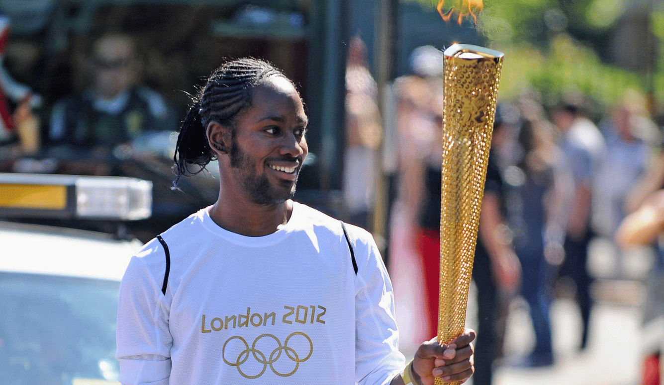 Young black man with braided hair and a short beard carrying the London 2012 Olympic torch while wearing a white London 2012 shirt outdoors.