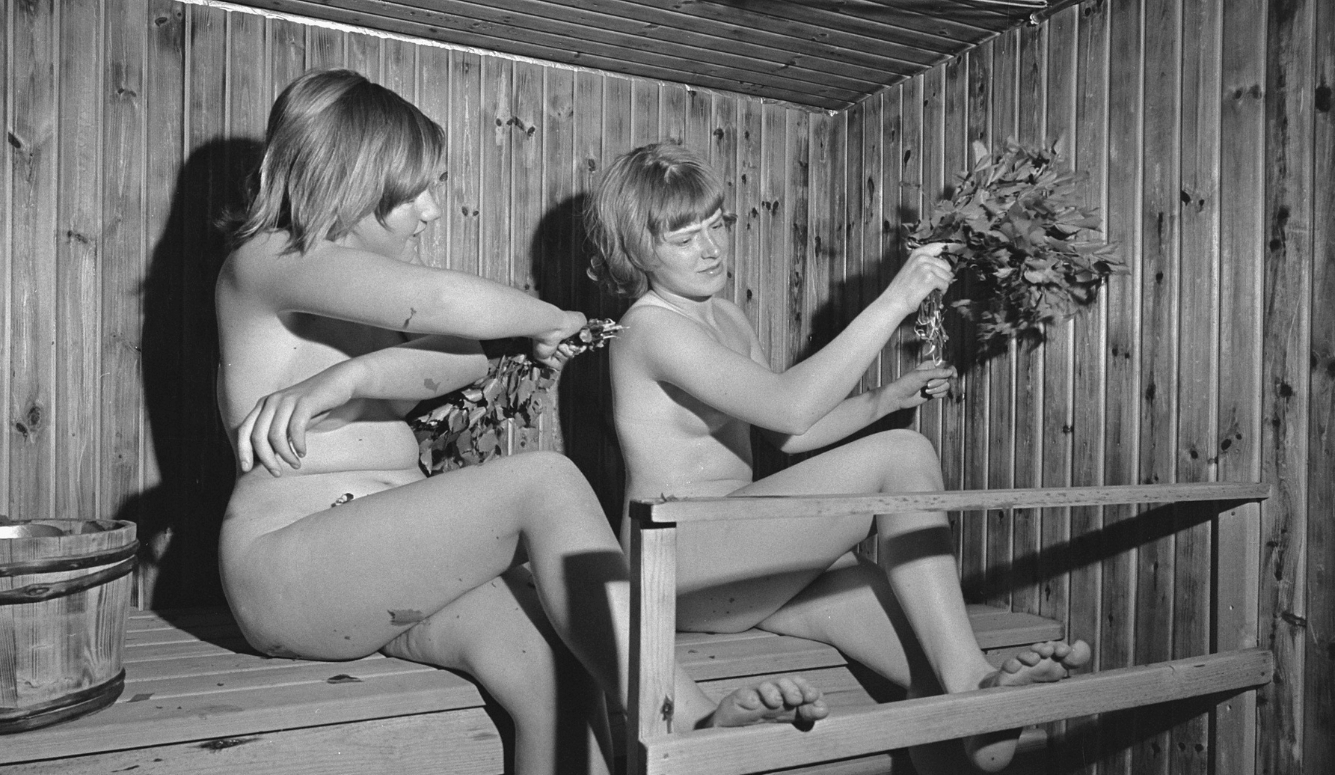 Two women naked in a sauna fanning themselves with birch branches.
