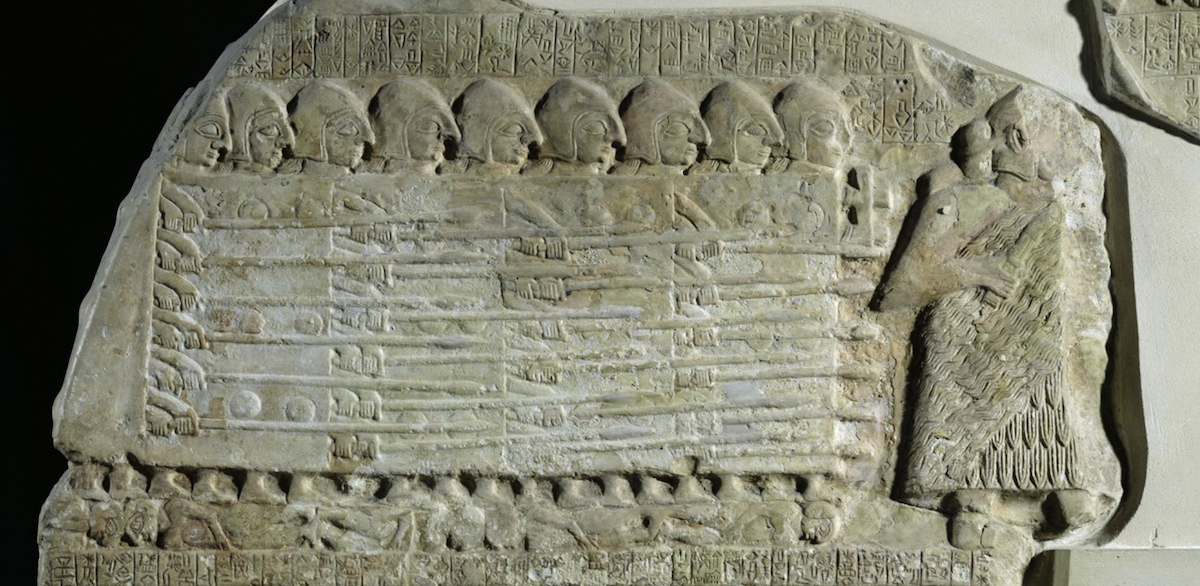 Ancient carved stone relief showing ranks of helmeted soldiers with spears led by a larger robed ruler.