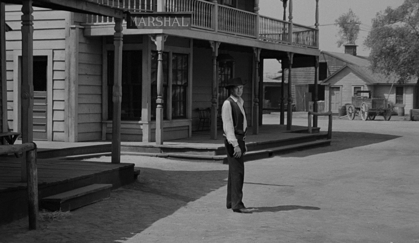 Gary Cooper as Marshal Will Kane stands alone in a dusty Western street outside the marshal's office in High Noon (1952).