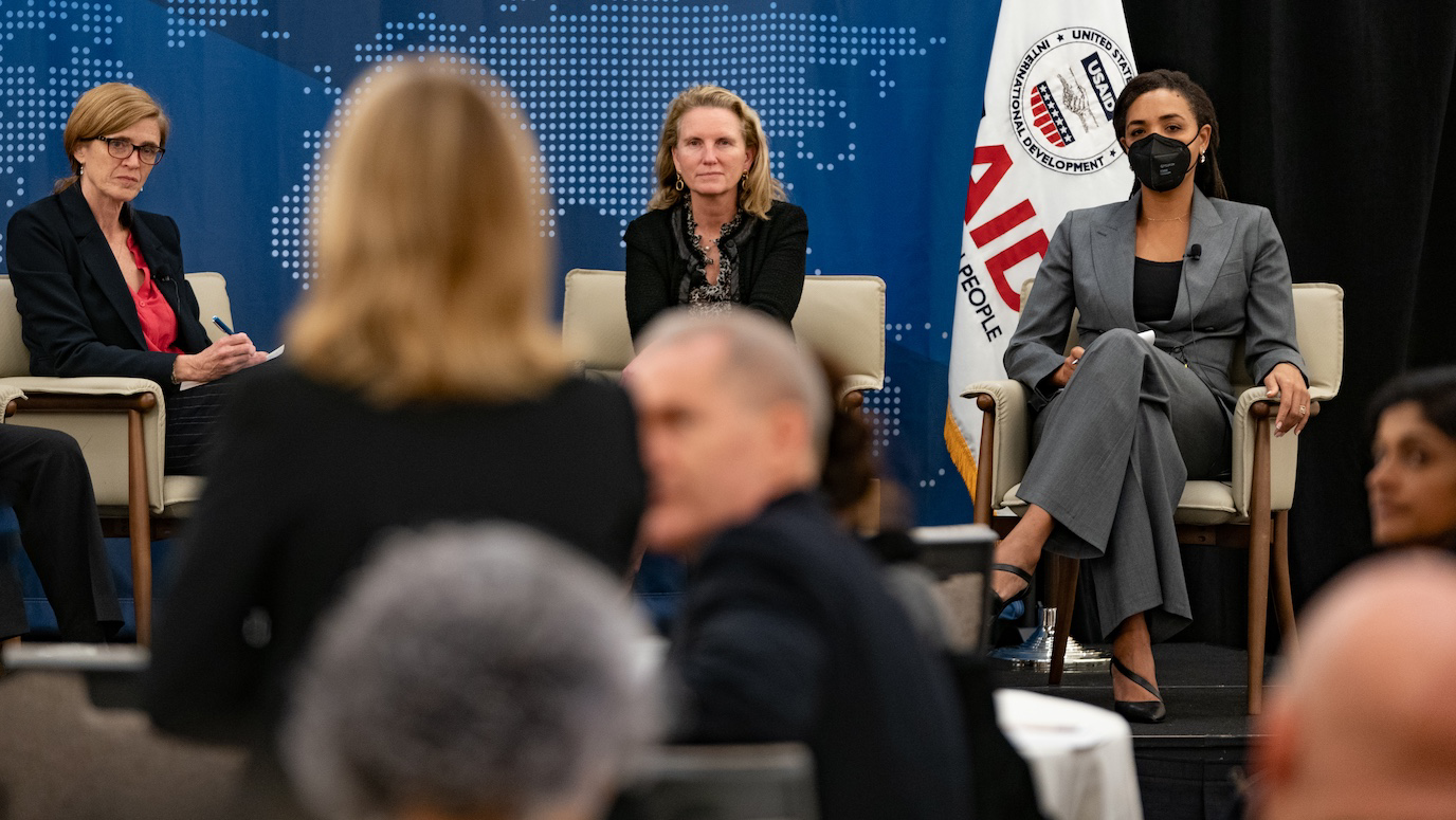 Three women seated on a panel at a USAID conference, with an audience in the foreground.