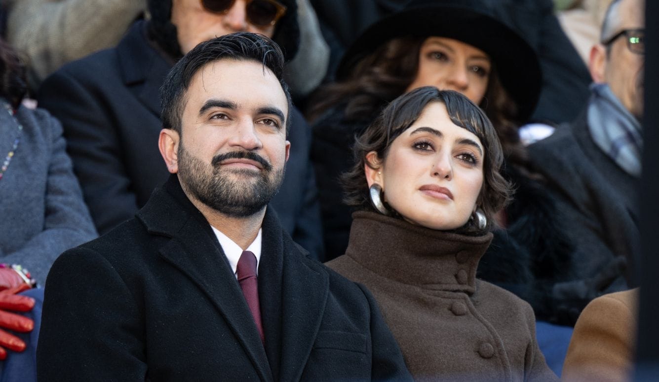 Zohran, a bearded man in a black coat and Ram in a brown coat seated among a crowd at an outdoor event.