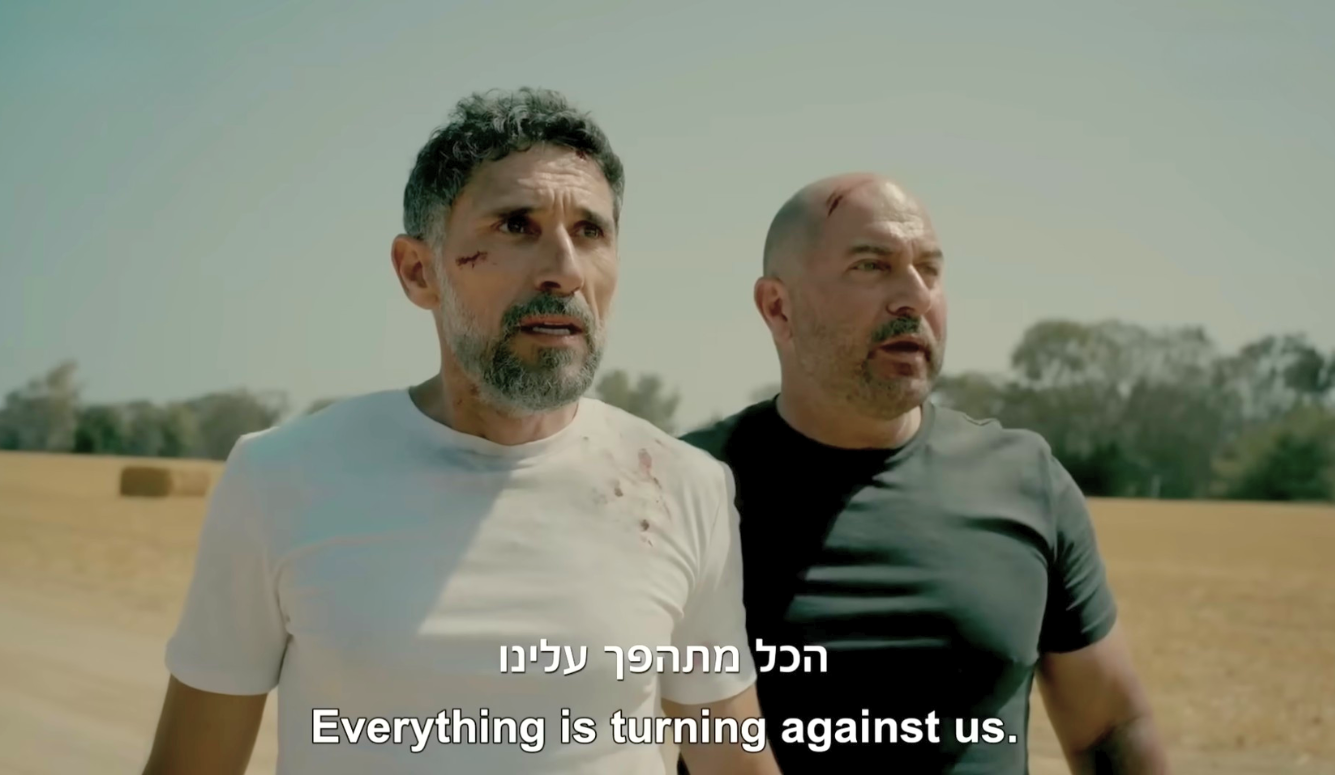 Two injured men in T-shirts stand in a field in a still from Fauda Season 5. The English caption in the photo says: “Everything is turning against us.”