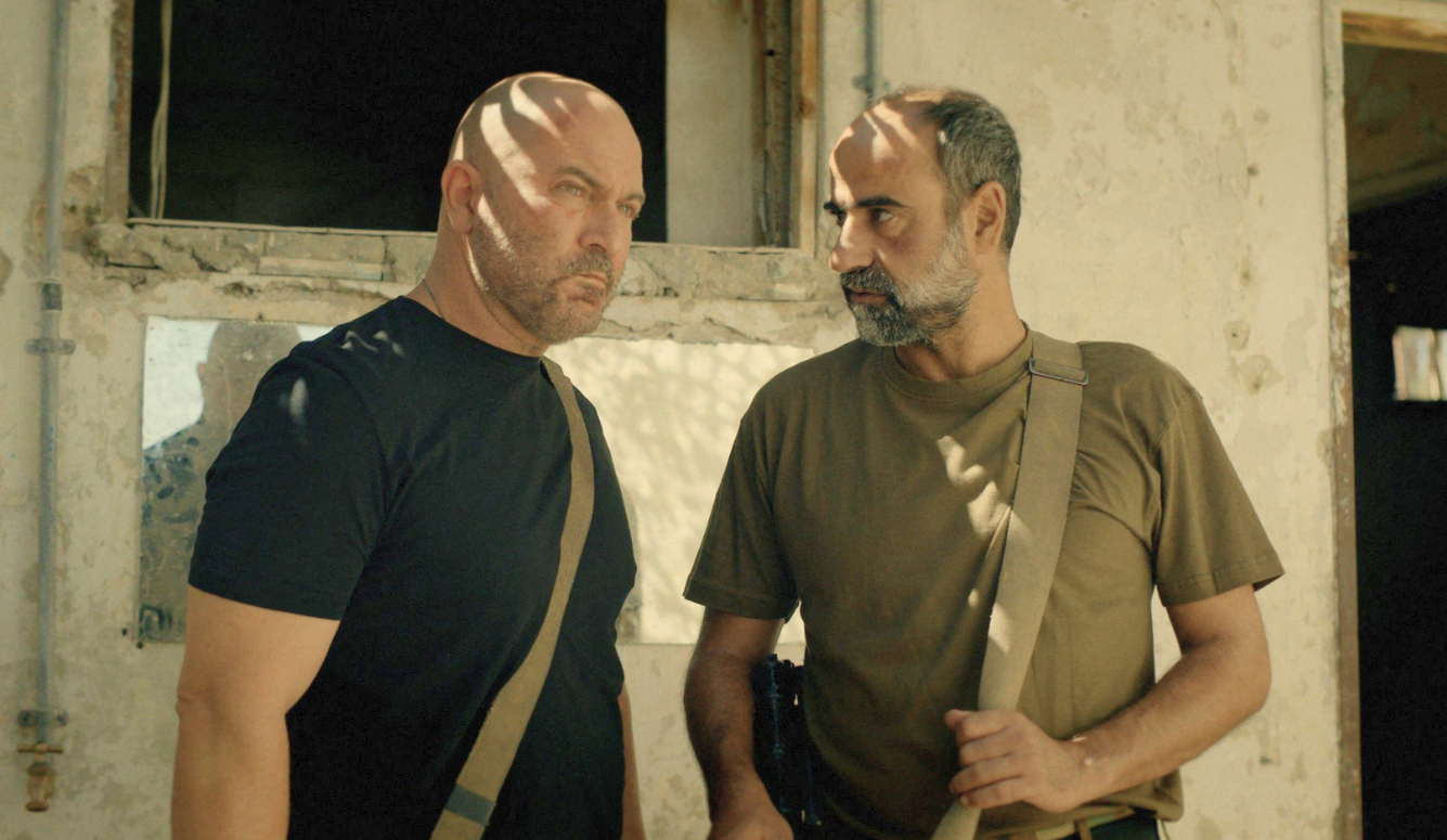 Two men in T-shirts and shoulder straps stand outside a weathered building in Fauda.