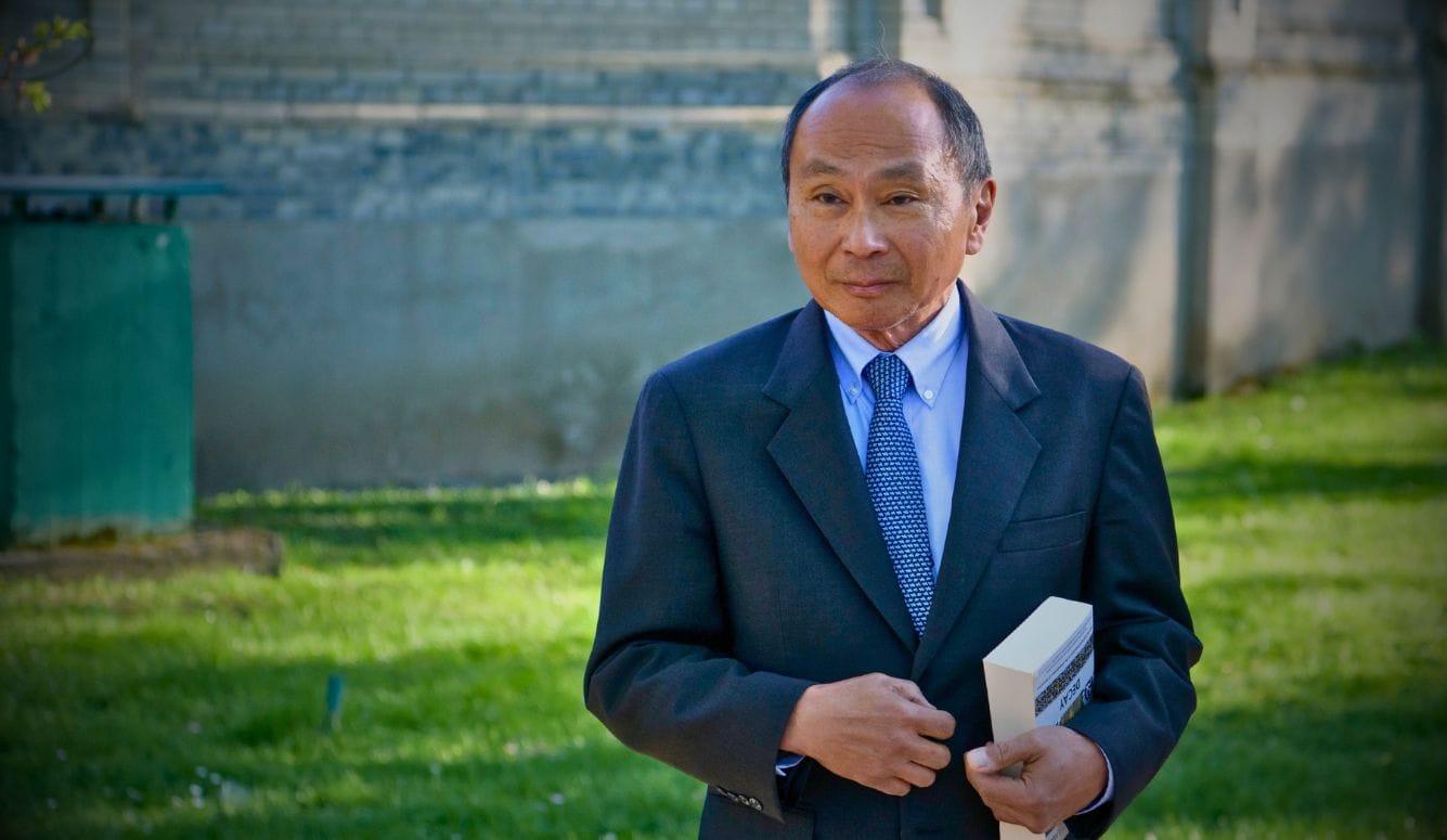Francis Fukuyama in a dark suit and patterned tie holding a book outdoors beside a stone building.