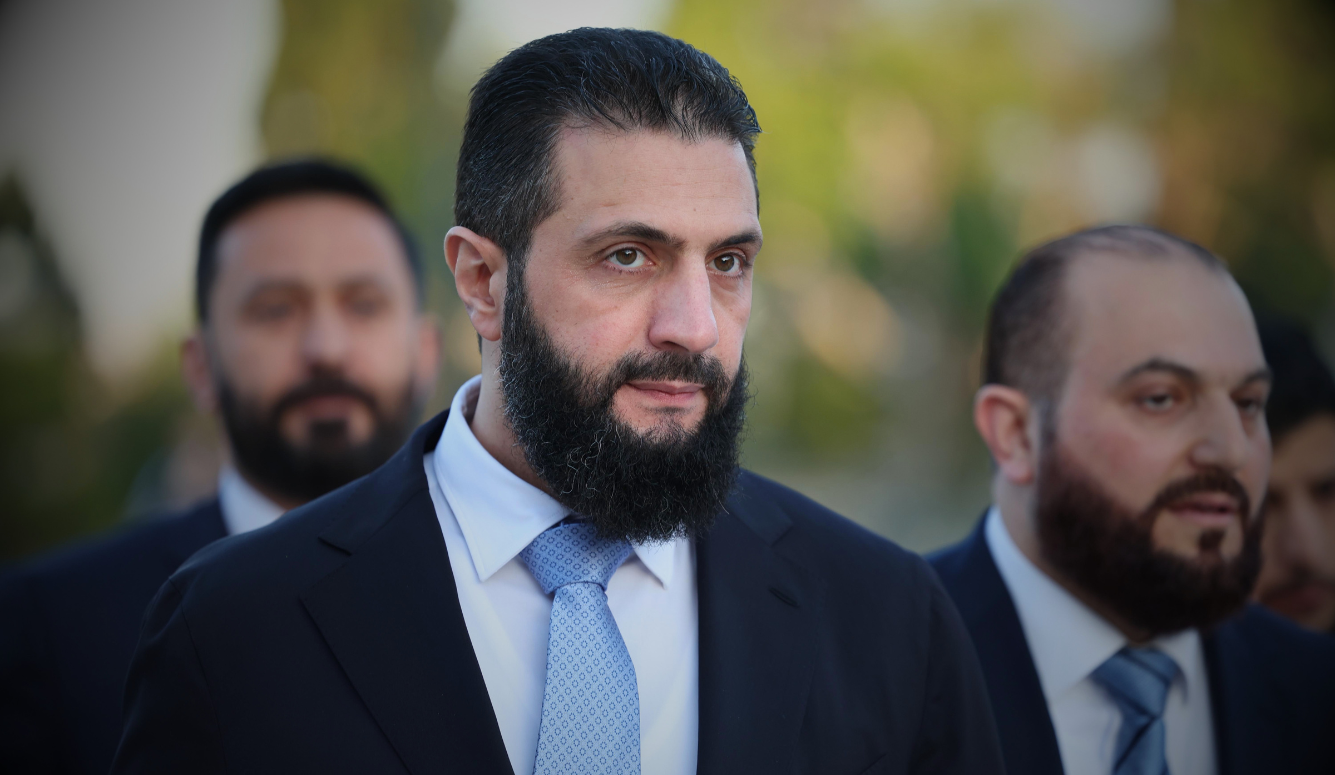 Syrian President Ahmed al-Sharaa in a suit and patterned tie, with two bearded men behind him outdoors