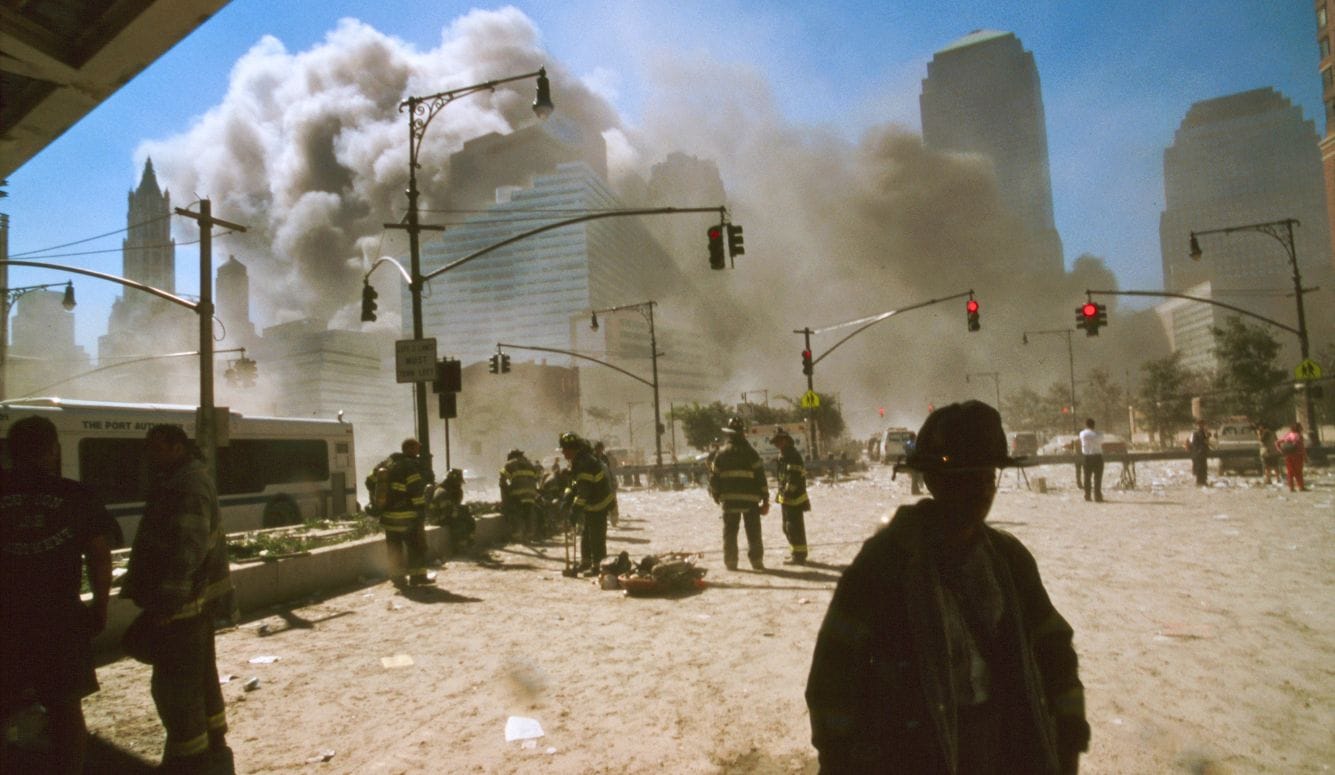 Firefighters and civilians in a dusty street as smoke rises among buildings in Lower Manhattan.