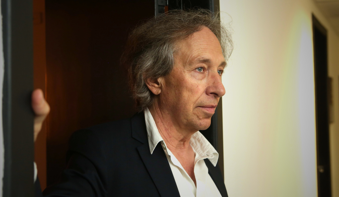 Older man with grey hair in a dark jacket and white shirt standing in a doorway.