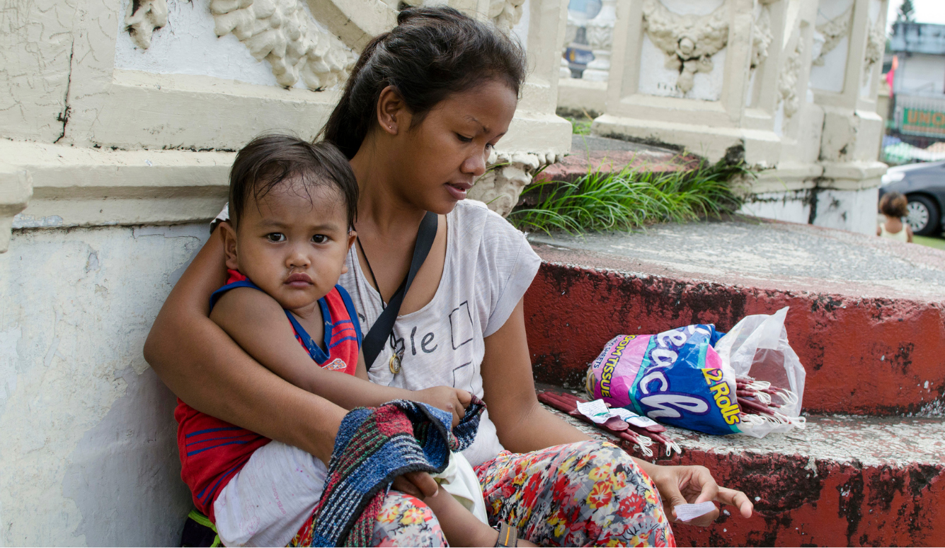 Woman holding a baby boy sits on church steps beside packets of candles for sale.