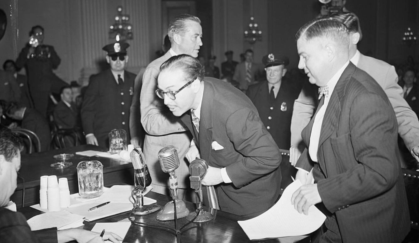 Man in glasses and suit leans towards microphones as police officers and officials stand behind him.