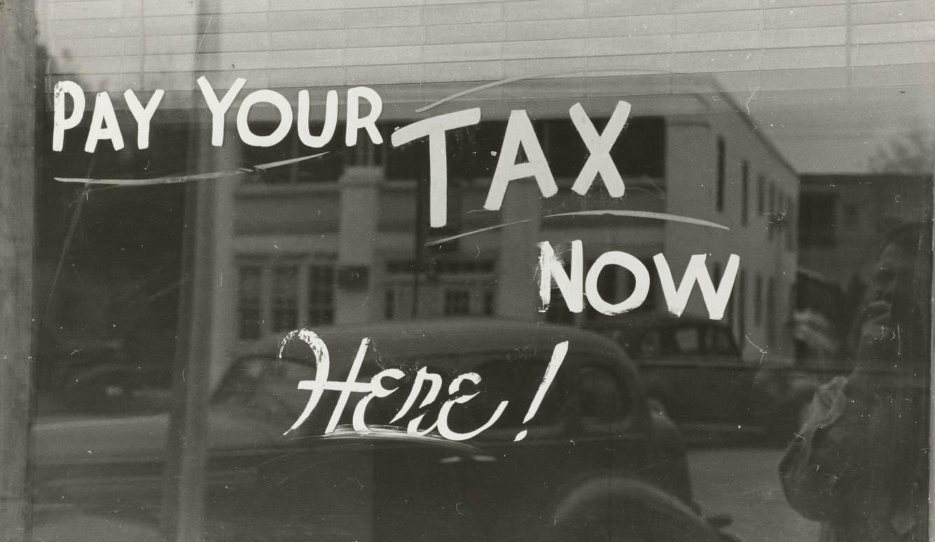 Black-and-white shop window with handwritten ‘Pay Your Tax Now Here'