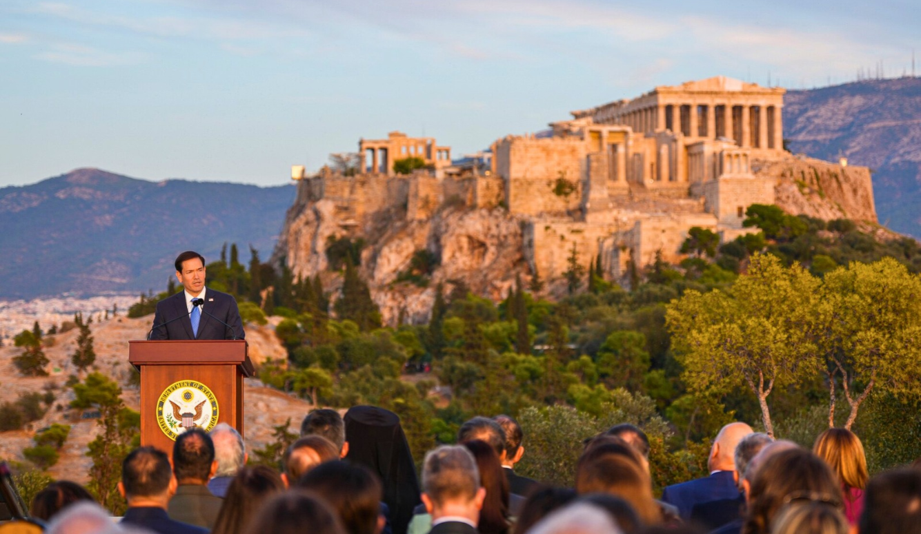 US Secretary of State Marco Rubio, wearing a navy suit and blue tie, speaks at a lectern before the Acropolis in Athens, Greece.