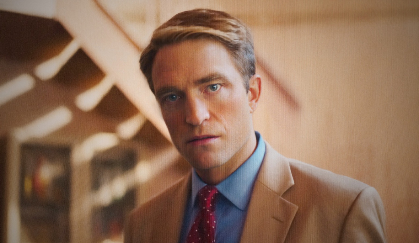 Robert Pattinson in a tan suit, blue shirt and red patterned tie, standing in a warmly lit interior.