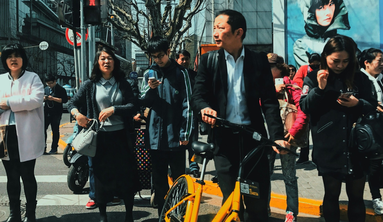 A man in a dark suit walks beside an orange bicycle on a busy Shanghai street, surrounded by pedestrians using mobile phones.