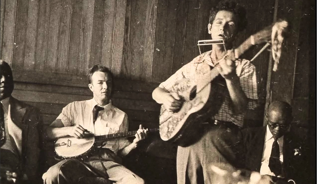 Woody Guthrie plays guitar and harmonica while Pete Seeger plays banjo in a wood-panelled room, accompanied by two seated men.