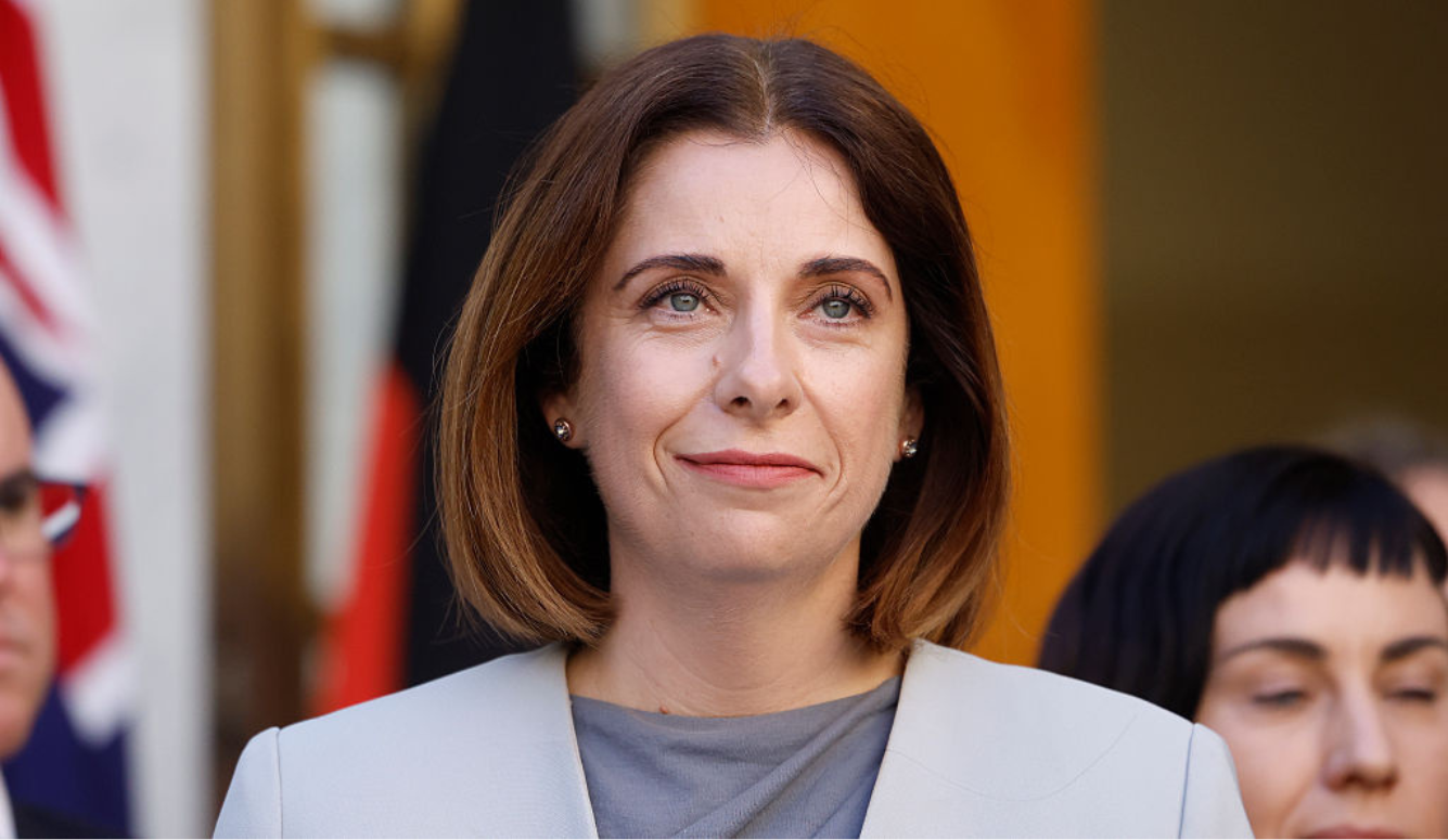 Anika Wells is a white middle-ag woman with a brown bob. She smiles in front of the Australian flag and wears a grey blazer. 
