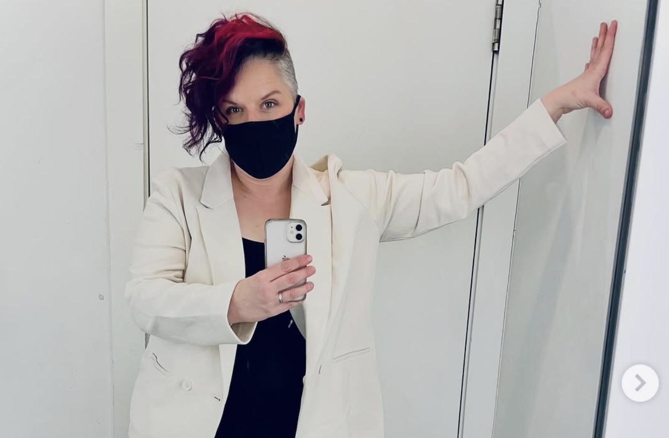 Rowan Jetté Knox takes a selfie in the mirror. She has bright pink and purple hair, which is cut short and shaved on one side. She wears a black COVID mask. 